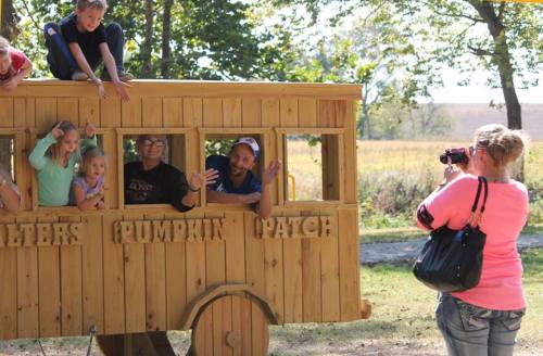 Children play on a wooden bus at a pumpkin patch while an adult takes their photo. Trees and fields are in the background.