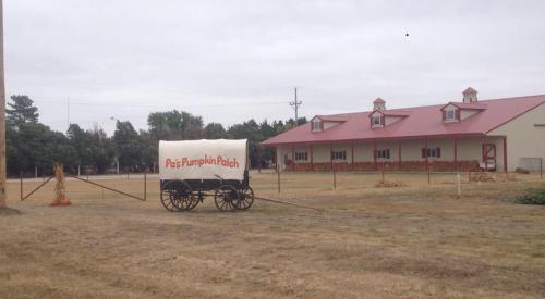 A rustic wagon with a white cover sits in a field near a barn with a red roof, surrounded by dry grass.