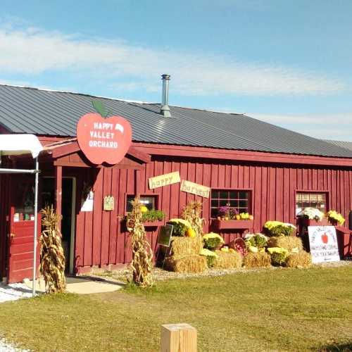 A red barn-style building with a sign reading "Happy Valley Orchard," surrounded by hay bales and colorful flowers.