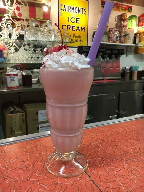 A tall glass of pink milkshake topped with whipped cream and a cherry, served at a retro-style diner.