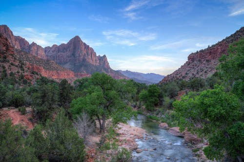 A serene landscape featuring a river winding through lush greenery, surrounded by towering red rock mountains under a blue sky.