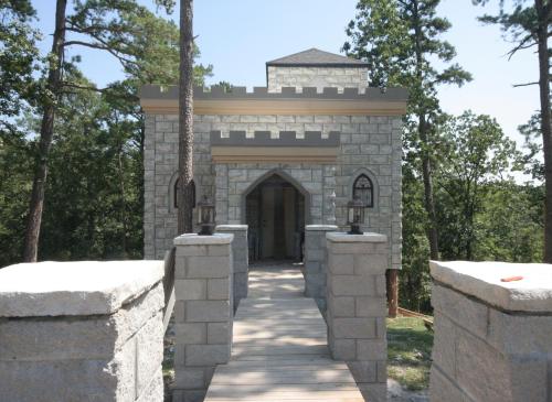 A stone castle with a wooden bridge leading to its entrance, surrounded by trees in a sunny outdoor setting.