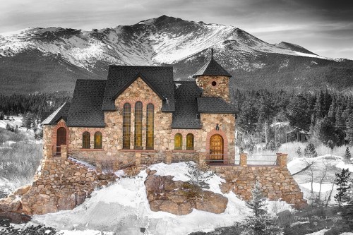 A stone house with large windows sits on a rocky outcrop, surrounded by snow and mountains in the background.