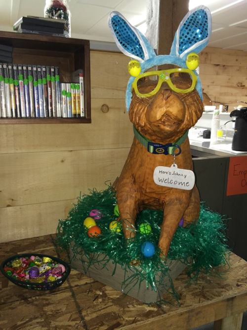 A wooden dog statue wearing bunny ears and sunglasses, surrounded by colorful Easter eggs and green grass.