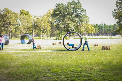 Children playing in a grassy field with large circular tubes and hay bales, enjoying outdoor activities.