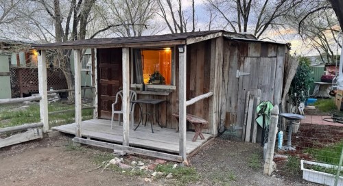 A rustic wooden cabin with a porch, surrounded by trees and a garden area, at dusk.
