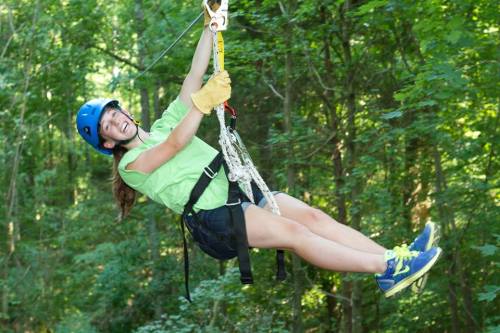 A girl in a green shirt and helmet zip-lining through a lush forest, smiling as she enjoys the adventure.