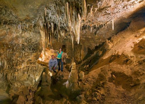 Two people explore a cave with stalactites and stalagmites, illuminated by soft lighting.