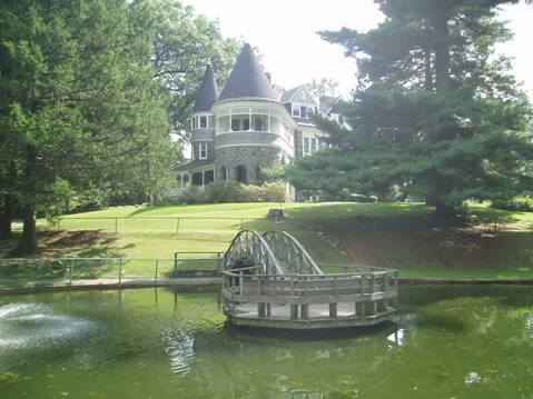 A large, historic mansion with turrets overlooks a pond, featuring a small wooden bridge and lush greenery.