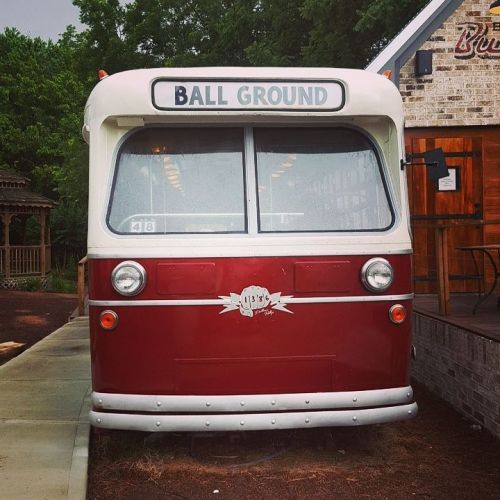 A vintage red bus with "BALL GROUND" on the front, parked near a rustic building and surrounded by greenery.