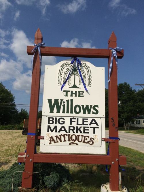 Sign for "The Willows Big Flea Market Antiques" with decorative blue ribbons, set against a clear blue sky.