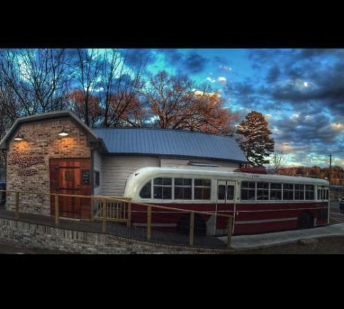 A vintage bus converted into a building, set against a colorful sky and trees, with a wooden deck in front.
