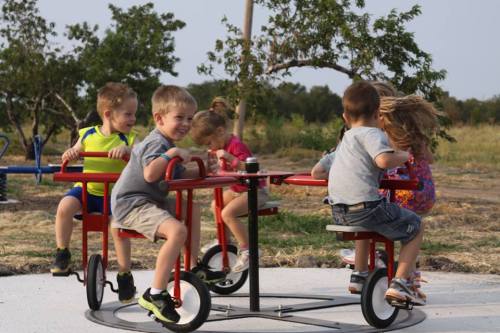 Five children play on a spinning playground merry-go-round, laughing and enjoying a sunny day outdoors.