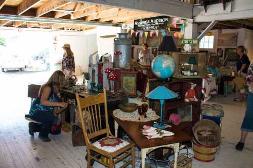 A vintage market scene with various antiques, furniture, and shoppers browsing in a rustic indoor space.