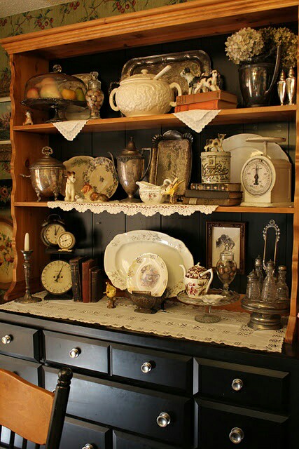 A vintage hutch displaying various antique dishes, silverware, clocks, and decorative items on lace doilies.
