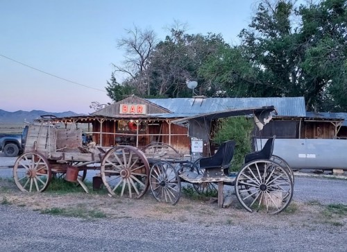 Old wooden wagons outside a rustic bar with a neon "BAR" sign, set against a backdrop of trees and mountains.
