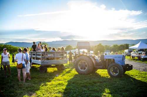 A blue tractor with a wooden trailer parked in a field, with people gathered around under a bright sunset.