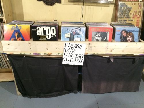 A wooden display with vinyl records and a sign that says, "Please take one tag to cash."