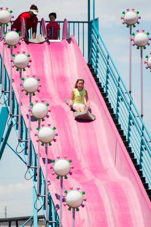 A girl slides down a pink slide at a fair, while two children play at the top. Colorful lights line the slide.