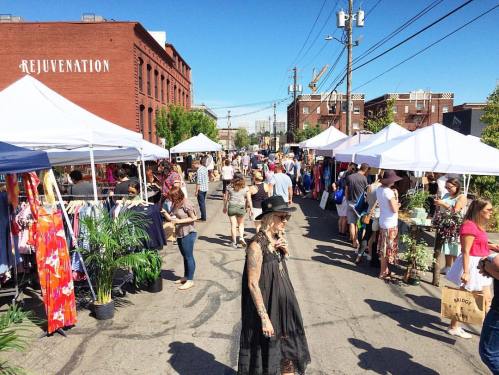 A bustling outdoor market with vendors under white tents, shoppers browsing colorful items on a sunny day.