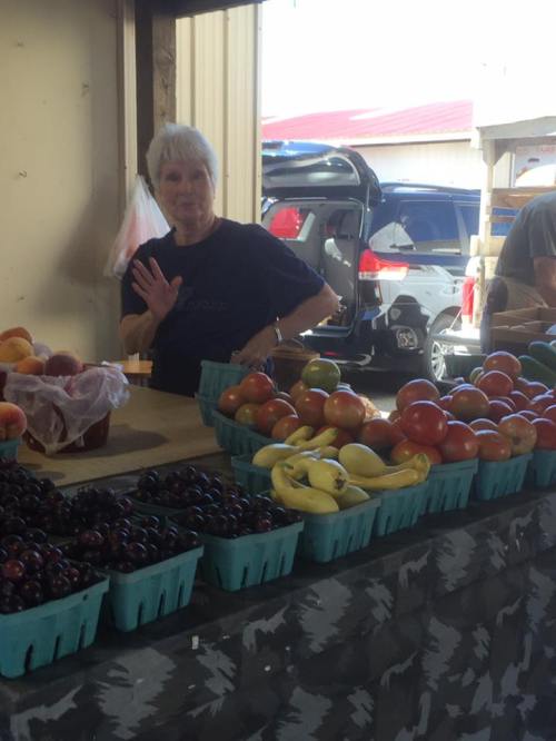 A woman waves from behind a table filled with fresh fruits at a market, with a car visible in the background.