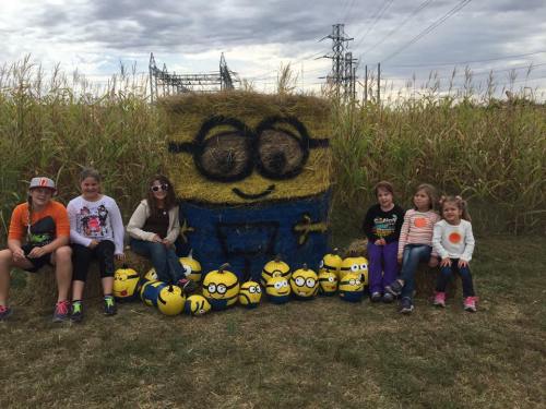 Children sit in front of a large haystack shaped like a Minion, surrounded by smaller Minion decorations in a field.