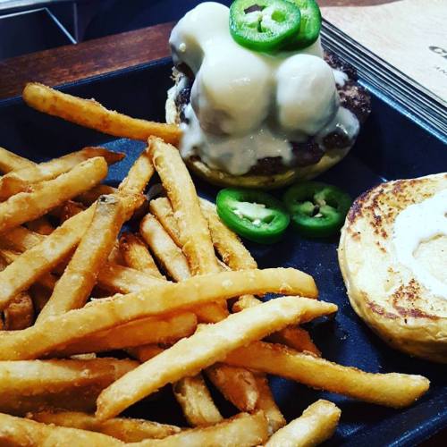 A plate with a burger topped with jalapeños and two white sauces, accompanied by a side of golden fries.