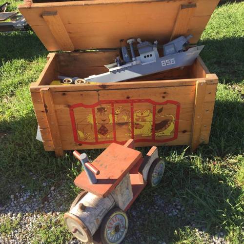 A wooden toy box with a model boat inside, next to a small wooden tractor on grass.