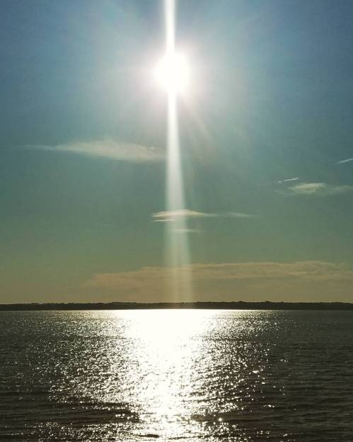 Bright sun reflecting on calm water, with a clear blue sky and distant land on the horizon.