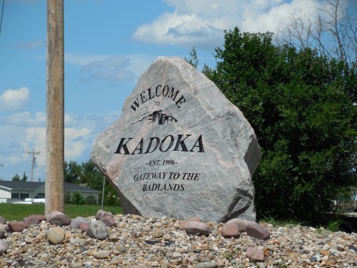 A large stone sign reading "Welcome to Kadoka - Est. 1906 - Gateway to the Badlands," surrounded by rocks and greenery.