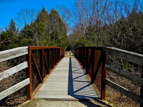 A wooden bridge spans a path through a wooded area, with clear blue skies above and shadows cast on the ground.