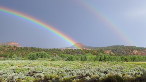 A vibrant rainbow arcs over green hills and a sagebrush landscape under a cloudy sky.