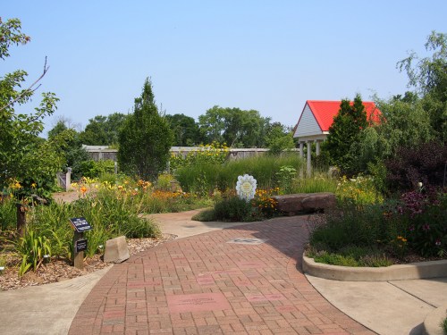 A winding path through a vibrant garden with flowers, greenery, and a small building with a red roof in the background.