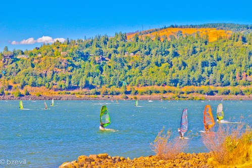 Colorful windsurfers glide across a vibrant blue river, surrounded by lush green hills under a clear sky.