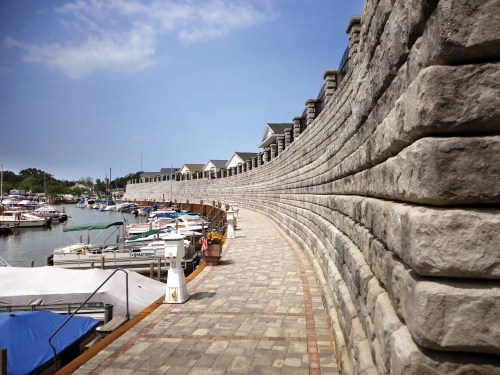 A curved stone wall lines a marina with boats docked along the water under a clear blue sky.