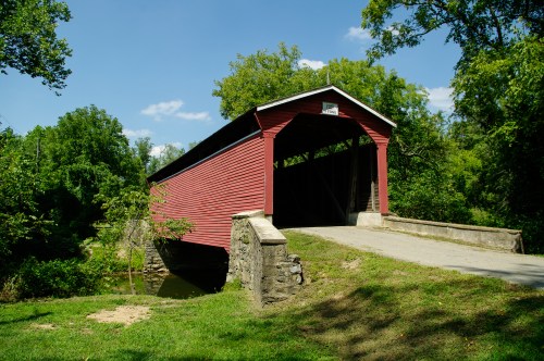 A red covered bridge spans a small stream, surrounded by lush green trees and a clear blue sky.