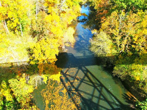 Aerial view of a river surrounded by vibrant autumn foliage, with a shadow of a bridge cast over the water.