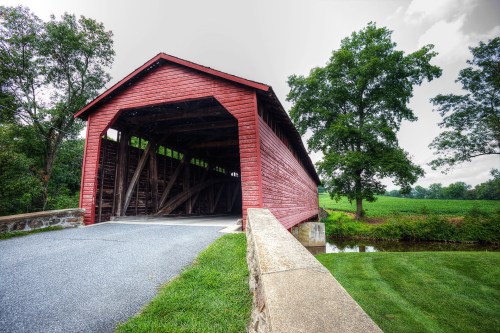 A red covered bridge stands over a gravel path, surrounded by green fields and trees under a cloudy sky.
