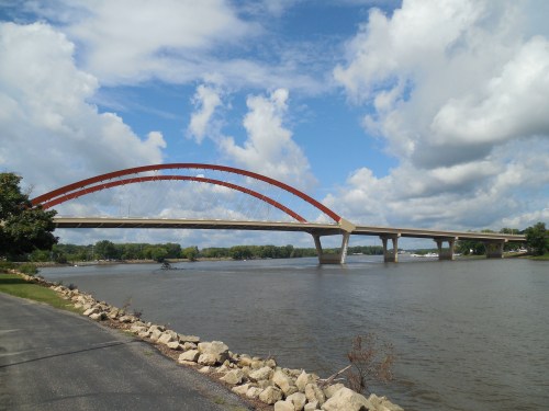 A modern bridge with a red arch spans a river under a partly cloudy sky, surrounded by greenery and a pathway.
