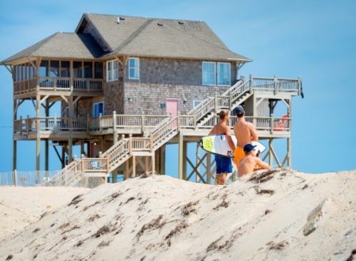 Three young men on a beach with a large house in the background, one holding a surfboard.