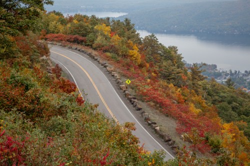 Winding road through vibrant autumn foliage overlooking a lake and distant hills.