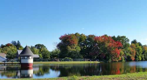 A serene lake scene with a gazebo, colorful autumn trees, and a clear blue sky reflecting on the water.