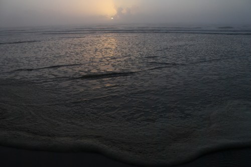 A misty beach at sunset, with gentle waves and a reflective surface, creating a serene atmosphere.