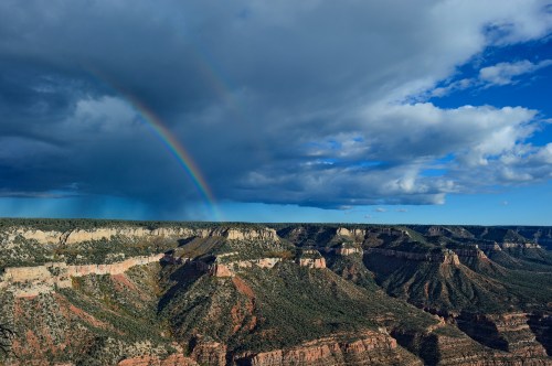 A vibrant rainbow arcs over the Grand Canyon, framed by dramatic clouds and rugged canyon walls.