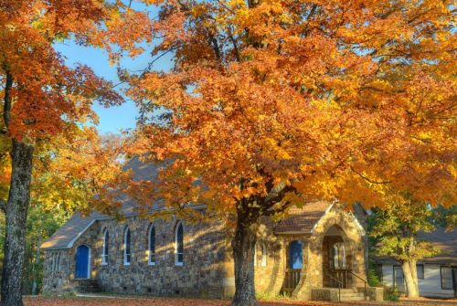 A stone church surrounded by vibrant autumn trees with orange and yellow leaves under a clear blue sky.