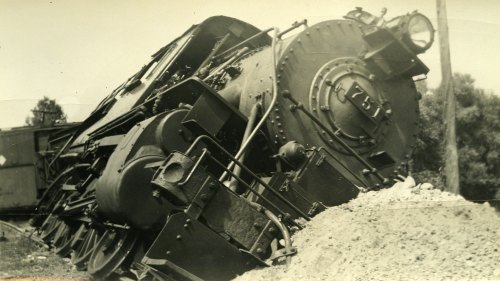 A vintage black and white photo of a derailed steam locomotive resting on its side near a pile of gravel.