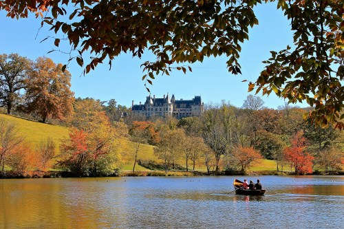 A scenic view of a lake with a boat and colorful autumn trees, featuring a grand mansion in the background.