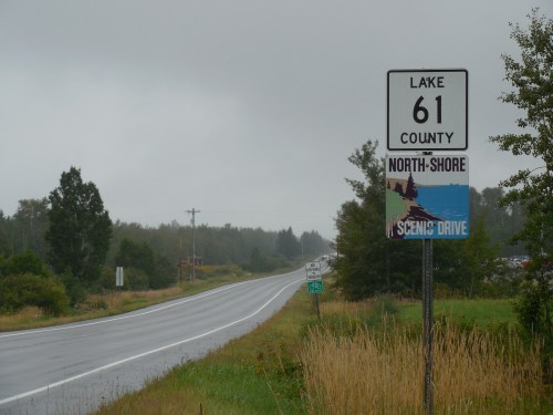 A road sign for County 61, indicating North Shore Scenic Drive, with a cloudy sky and a wet road in the background.