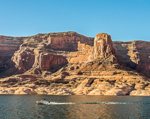 A boat glides across a calm lake, with towering red rock formations and clear blue skies in the background.