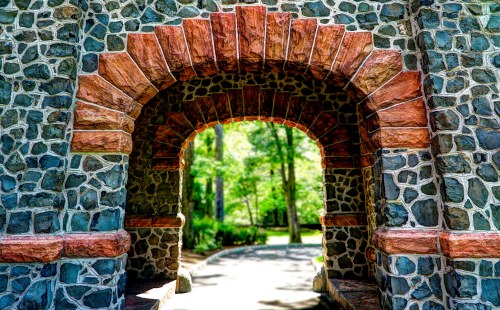 A stone archway with red accents frames a view of a lush, green park path beyond.